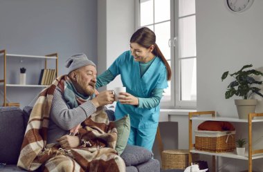Caregiver giving a cup of hot tea to a frozen senior man wrapped in a blanket in hat at home. Nursing home service and patient support. Cheerful brunette nurse helping an elderly man and smiling.