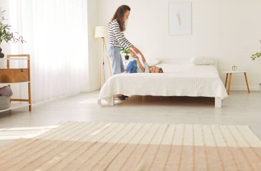 Mother and child spending time at home. Happy mom holding hands of her little kid daughter who is lying on a comfortable bed in a light spacious bedroom. Family, leisure concept