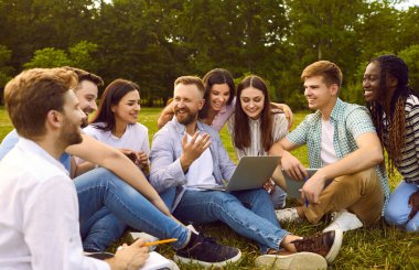 Group of happy, cheerful, diverse, multiethnic student friends sitting on green grass outside, studying information for project together, discussing something, using online tools on modern laptop PC