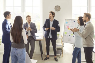 Cheerful business team applauding colleague during presentation near whiteboard in modern office. Group of smiling professionals clapping hands and celebrating success after meeting or training.