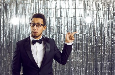 Surprised man in classic black tuxedo points his index finger sideways at advertising copy space. Portrait of young dark-skinned man on shiny silver background. Concept of show, event, party or