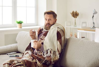 Portrait of sick young man drink hot tea measure high temperature struggle with corona virus on lockdown at home. Unhealthy male suffer from covid-19, have flu or fever. Healthcare concept.