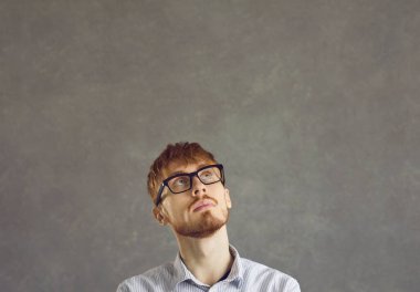 Thinking casual male hipster nerd man freelance programmer looking up feeling skeptic doubtful. Thoughtful suspicious guy in glasses standing over grey studio copy space wall. Contemplation