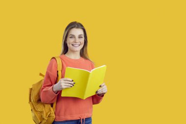 Portrait of smiling female student with backpack and textbook isolated on yellow background. Happy girl in orange sweatshirt holds open notebook and looks at camera standing near copy space. Banner,