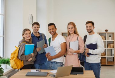 Portrait of happy multiracial students with backpacks and books pose together in school classroom. Group of smiling diverse young people learners in college or university. Education and courses.