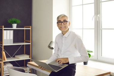 Portrait of happy successful businessman with laptop at work. Senior business man standing by office desk, holding portable notebook computer, looking at camera and smiling