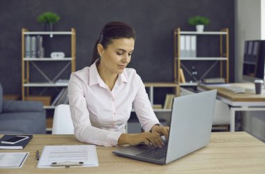 Smiling young woman typing on a laptop while sitting at her workplace in the office. Young European woman doing her office work, documents, smartphone, notebook and pen lie on the desktop.