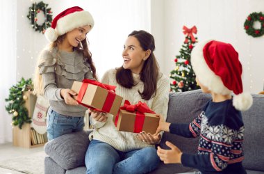 Happy joyful young mother gets unexpected presents from her loving children on Xmas Day. Woman with pleasantly surprised face receives greetings from her young son and daughter on Christmas morning.