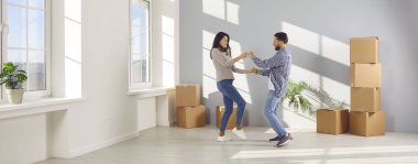 Happy young couple celebrating moving day with cardboard boxes in background smiling and dancing with in new appartment. Family having fun. Relocating, real estate, mortgage concept. Banner.