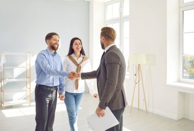 Happy couple buying house. Man and woman standing in modern white light living room and shaking hands with real estate agent. Young family buy property, make deal and exchange handshake with relator