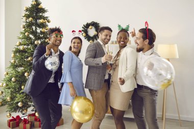 Happy friends having fun at Christmas party. Group of cheerful young diverse people in suits, dresses and funny headbands enjoying music and dancing in room with decorated Xmas tree