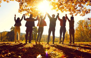 Unity and friendship. Group of friends holding hands raise them up while standing in sunlight in autumn park. Rear view of people in casual clothes standing with backs to camera and holding hands.