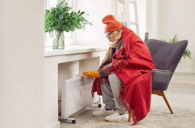 Oturma odasında sandalyeye oturmuş, battaniyeye sarılmış ve elektrikli ısıtıcıda ısınmaya çalışan yaşlı bir adamın portresi. Evde donan olgun bir adam. Isıtma sorunları konsepti.