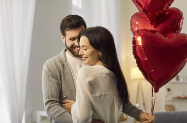 Happy couple celebrating Valentines Day at home, embracing affectionately with red heart-shaped balloons in the background. Love, affection, family bonding, and romantic holiday joy.