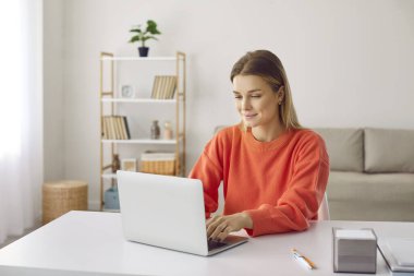 Ambitious young woman working remotely or distance learning at home using laptop. Smiling caucasian girl typing on laptop while sitting at desk in home office. Freelance or studying concept.