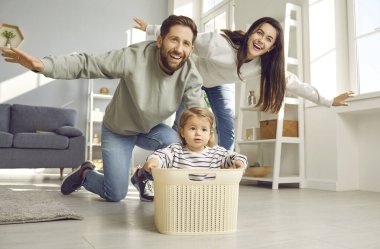 Dad, mom and toddler baby having fun playing at home. Cheerful parents playing together flying like airplane, spreading his arms to sides, cute kid sitting in plasatic box in living room at home