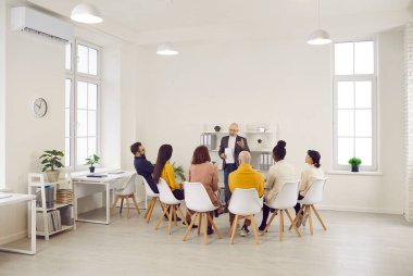 Old businessman lead head meeting with diverse employees or workers in office. Mature grey-haired confident male coach or trainer make presentation for businesspeople. Business training.