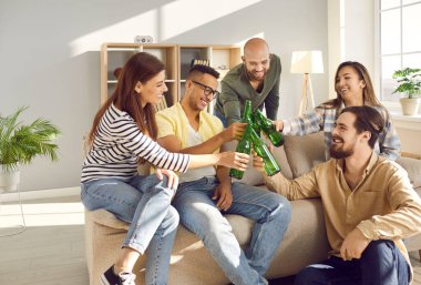 Smiling multiracial young people clink bottles relax at home together watching TV or football game. Happy diverse friends cheers with beer enjoy weekend indoors have fun rest indoors. Friendship.