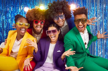 Portrait of funny group of happy young people dressed in bright costumes, voluminous curly wigs and colorful large glasses. Group of friends fooling around on the background of blue shiny wall.