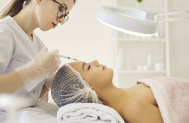 Güzellik uzmanı güzel bir kadının yüzüne kaş aralarında kozmetik enjeksiyon yapar. Estetik prosedürleri gençleştirirken müşterinin kanepeye uzanması. Yüz kırışıklıklarının düzeltilmesi kavramı.