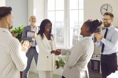 Two african american women shaking hands celebrating success, making deal, business achievement, signing contract or greeting new employee with group of people applauding in background in the office.