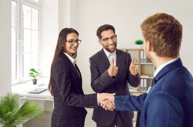 Professional business people make a deal, thank each other for cooperation and exchange handshakes. Happy woman shakes hands with man and her coworker shows thumbs up. Teamwork, collaboration concept