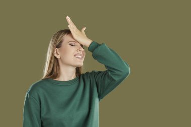 Disappointed embarrassed confused smiling blondy woman with long hair and closed eyes wearing green sweatshirt with facepalm gesture on khaki background with copy space. Sincere emotions concept.