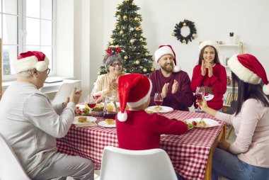 Joyful family having Christmas dinner meal. Happy mother, father, children and grandparents in red Santa caps sitting at festive table, enjoying delicious food, drinking juice and having fun together