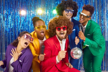 Group of cheerful, funny and creative young people having fun at karaoke party night. Multiracial people in colorful clothes, wigs and with disco ball sing into microphone next to shiny foil curtain.