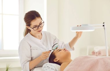 Dişi güzellik uzmanı elinde küçük bir büyüteçle derinin durumunu inceliyor. Bayan müşteri güzellik salonundaki resepsiyonda özel lambanın altında kanepede yatıyor. Güzellik konsepti.