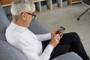 Concept of older adults using online dating apps or websites. Old bachelor searching for future wife. Senior man holding mobile phone in hand and looking at photo of beautiful young woman on display