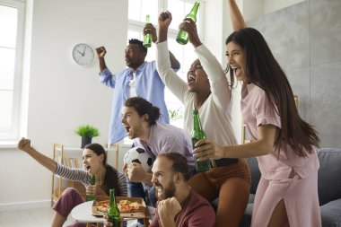 Group of young multiracial male and female football fans watching match on TV, drinking beer and supporting favourite team. Excited multiethnic friends shouting hoping favorite soccer team scores goal