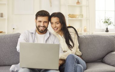 Happy laughing young couple watching laptop screen sofa sitting to enjoy funny movie, planning budget, reading good news in email, smiling looking at computer, checking finances at home together