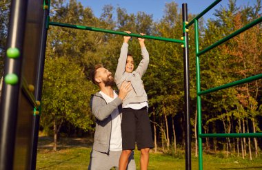 Genç bir baba ya da erkek koç, çocuk bir çocuğa dışarıdaki yaz parkında kendini yukarı çekmesine yardım ediyor. Sportif aile doğada spor egzersizleri yapıyor. Sağlıklı yaşam tarzı kavramı.