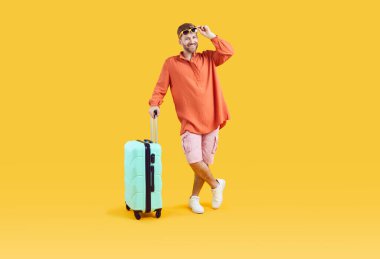 Studio portrait of happy male tourist. Full body length shot of joyful young man wearing comfortable summer clothes standing with his suitcase on yellow color background. Traveling, holiday concept