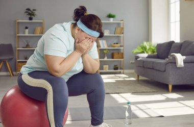 Portrait of a funny young fat overweight woman tired after fit sport exercises covering face with hands sitting on a fit ball at home. Workout, sport exercises and fitness concept.