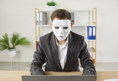 Anonymous man wearing suit and disguised in white face mask sitting at office desk and using laptop computer. Online business, anonymity, internet fraud, hacking, data theft, web privacy concept