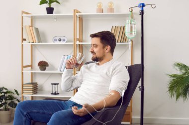 Dehydrated young man receiving intravenous vitamin therapy in hospital room. Male patient sitting in armchair attached to vitamin IV infusion drip in wellness center or beauty salon