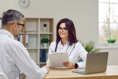 Friendly female doctor consults male patient filling out form during consultation. Smiling young woman in medical uniform sits with clipboard and listens to patient who sits with his back to camera.