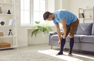 Young man standing by the sofa in the living room and putting elastic stretchable medical anti thrombosis or anti varicose sleeve socks or compression stockings on his tired legs