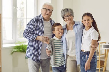 Portrait of a happy grandparents with their grandchildren boy and girl standing in the living room at home and looking cheerful at camera. Kids enjoying leisure time with grandma and grandpa.