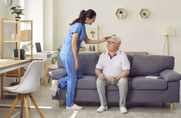 Friendly young female nurse combs hair of older man sitting on sofa in nursing home. Caregiver cares for elderly man in specialized institution for pensioners. Nursing and elderly care concept.