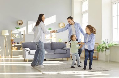 Funny active dancing family of young adult parents and cute small children together in modern living room interior, carefree little kids with mum, dad having fun laughing, enjoy leisure at home moving