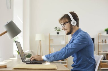 Young happy man in headphones typing on laptop and using internet web online services. Man relaxing and playing games or working remotely at home using modern technology and communication devices.