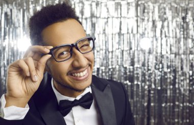 Close up of joyful dark skinned young man in suit on celebration blinks eye at camera. Guy with toothy smile wears glasses and suit with bow tie, posing on silver shiny background while at disco.