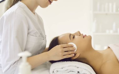 Female client enjoying cosmetic spa facial treatment at beauty parlour. Professional beautician cleaning young girls skin with cotton discs. Side profile view of womans head on soft white towel