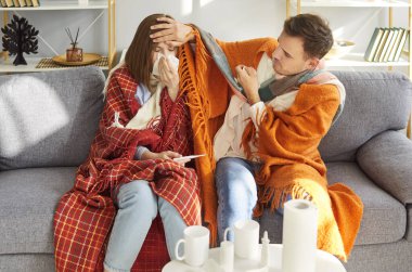Two sick young adults sitting on sofa under blankets in cozy living room. Caring man checking forehead of ill woman while she blowing her nose. Couple supporting each other during illness at home.