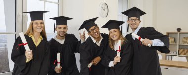 Happy students celebrating graduation. Cheerful mixed race multiethnic male and female university graduates in hats and robes standing in classroom, holding diploma scrolls and smiling at camera