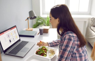 Woman eats food ordered at delivery service while sitting in her home office in front of laptop during lunch break. Side view of woman shopping online sitting at table with container of healthy food.