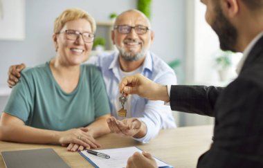 Bir emlakçı, satın almayı tamamladıktan sonra gülümseyen yaşlı bir çifte ev anahtarı veriyor. Ev sahipliğini ve başarılı bir kapanışı sembolize ediyor. Müvekkilleri olan emlakçı ofiste bir anlaşma yaptı..